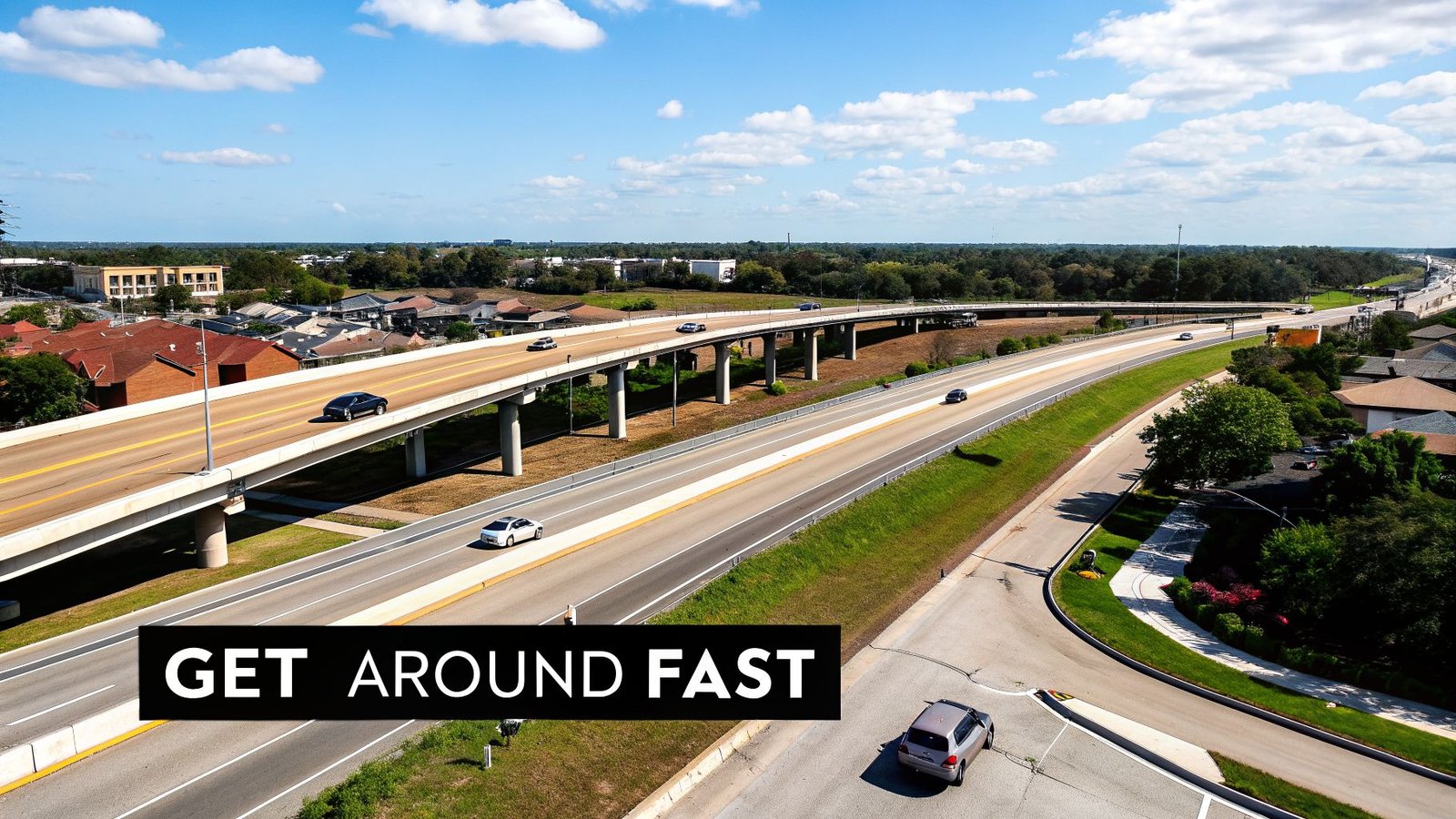 Aerial view of a busy multi-lane highway with an overpass, cars, houses, and green landscape under a blue sky.