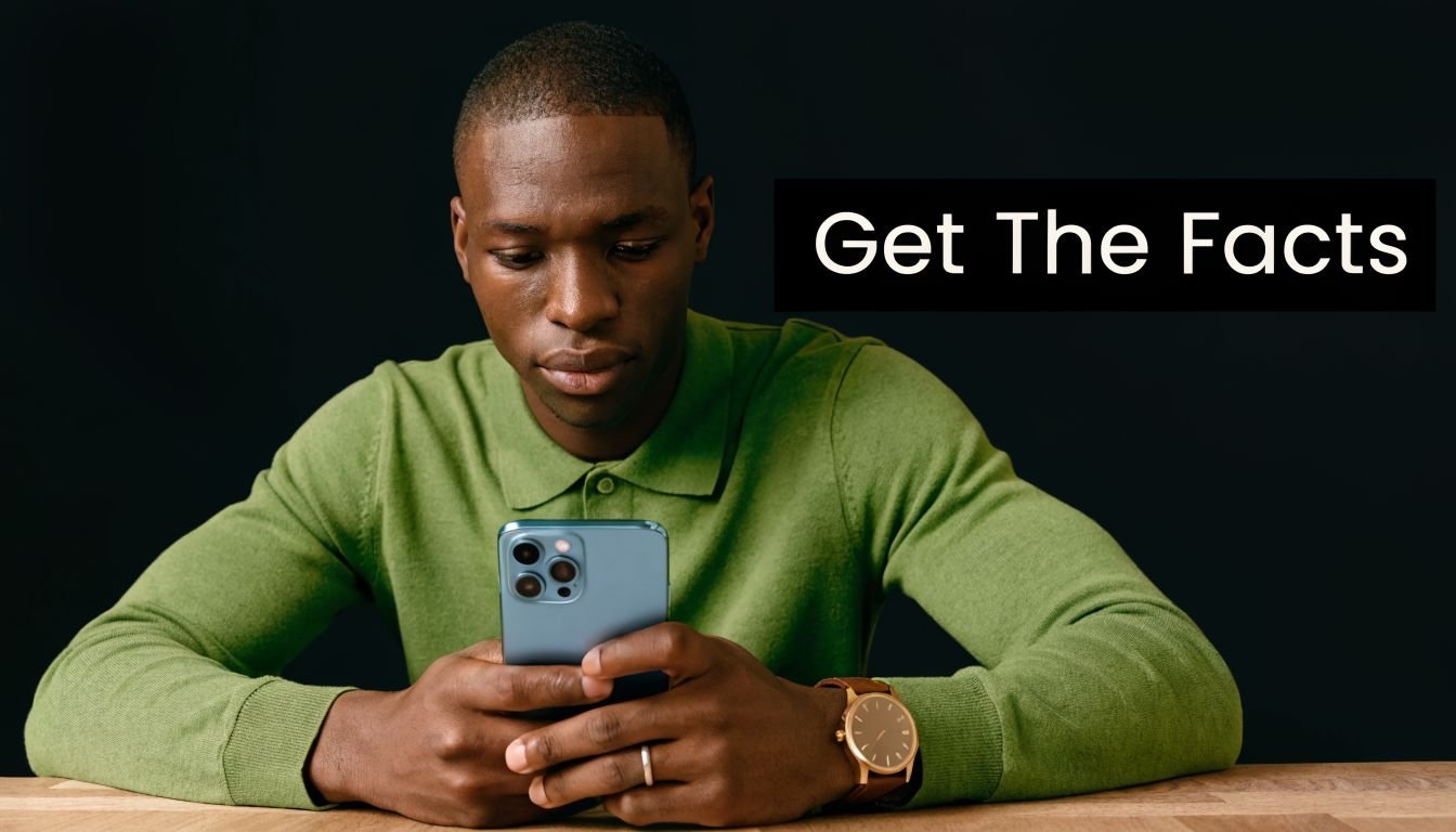 A young man wearing a green sweater looking down at his smartphone while seated at a table.