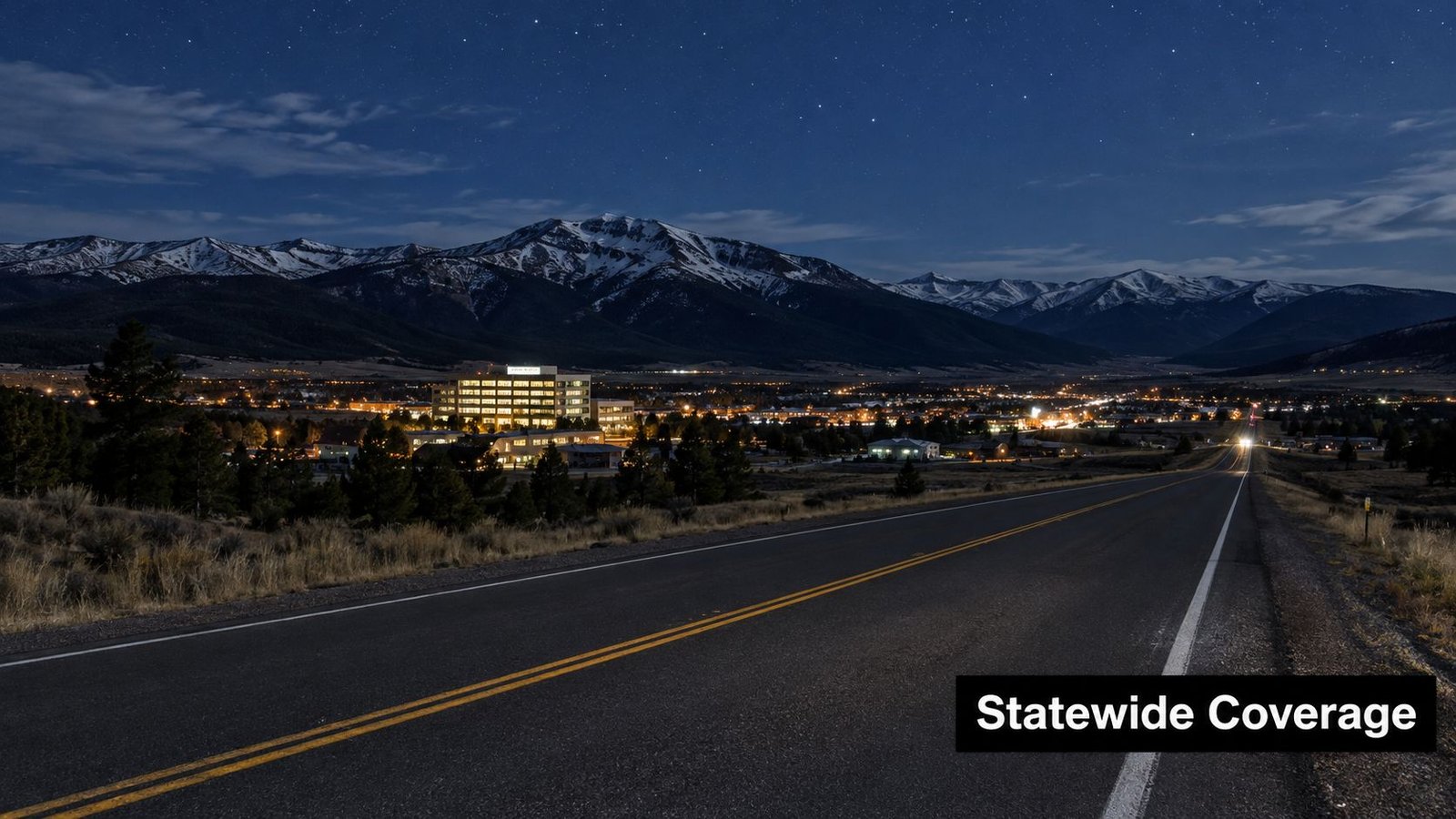 A scenic night view of a brightly lit town nestled beneath snow-capped mountain ranges under stars.